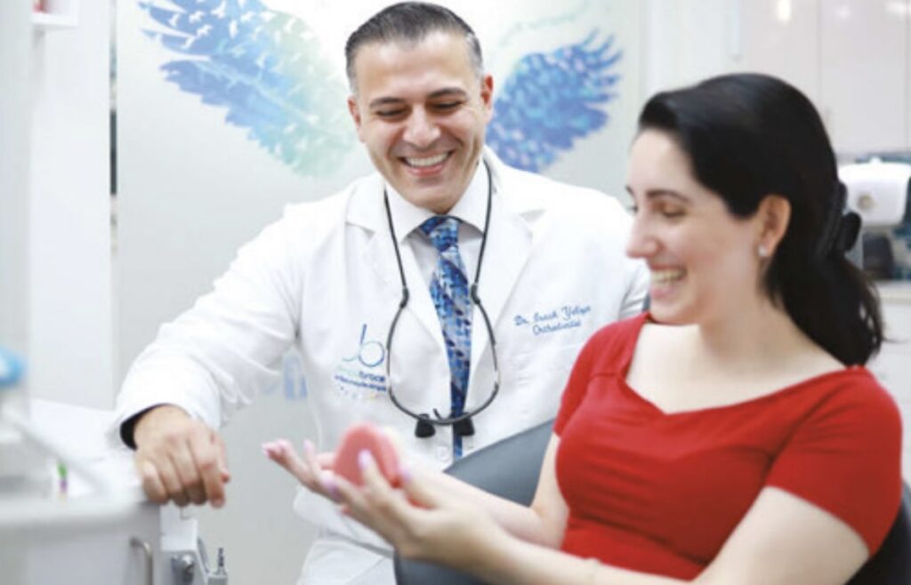 A dentist in a white coat smiles while talking to a female patient in a red top, who is sitting in a dental chair and looking at a dental model she holds. The background features a blue pattern on the wall.