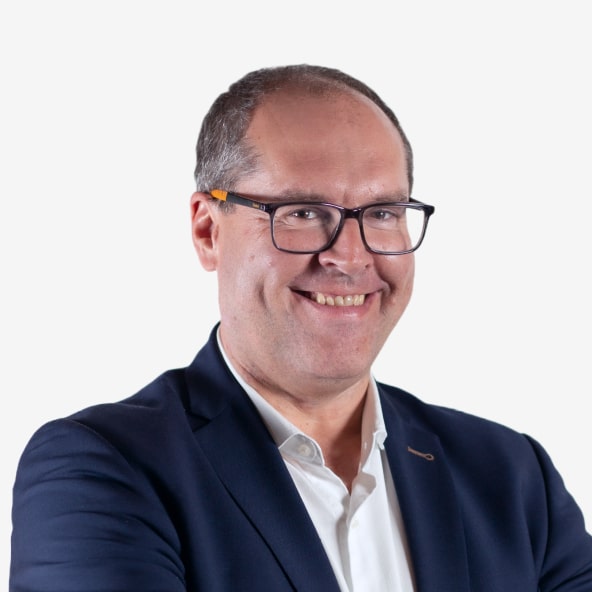 A man with short brown hair and glasses, smiling while wearing a navy blue suit jacket and white dress shirt, standing against a plain white background.