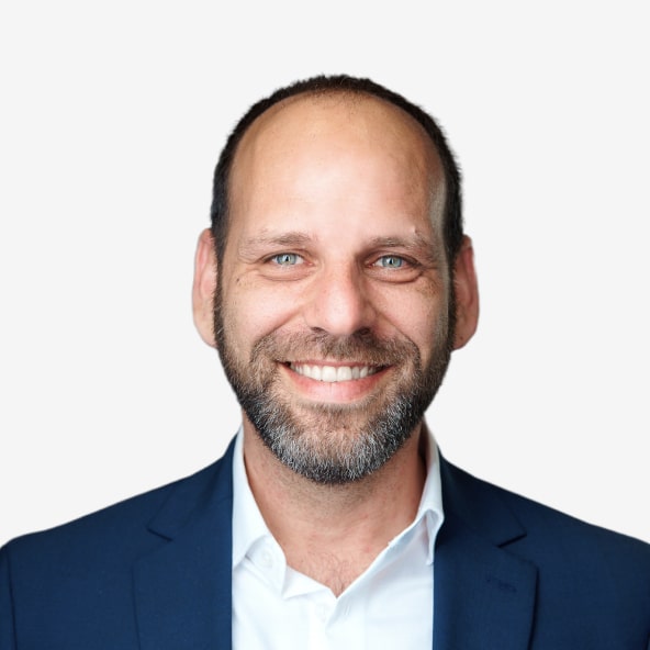 Smiling man with a beard and short hair wearing a navy blue suit jacket and white shirt, posing against a plain white background.