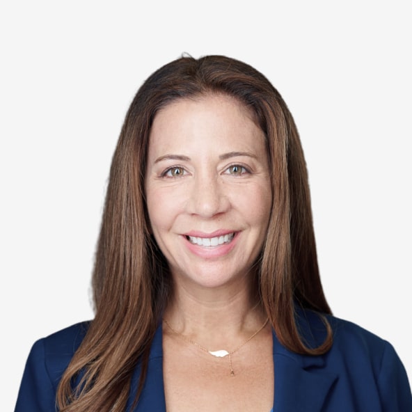 A woman with long brown hair, wearing a blue blazer and a delicate necklace, smiles at the camera against a plain light background.