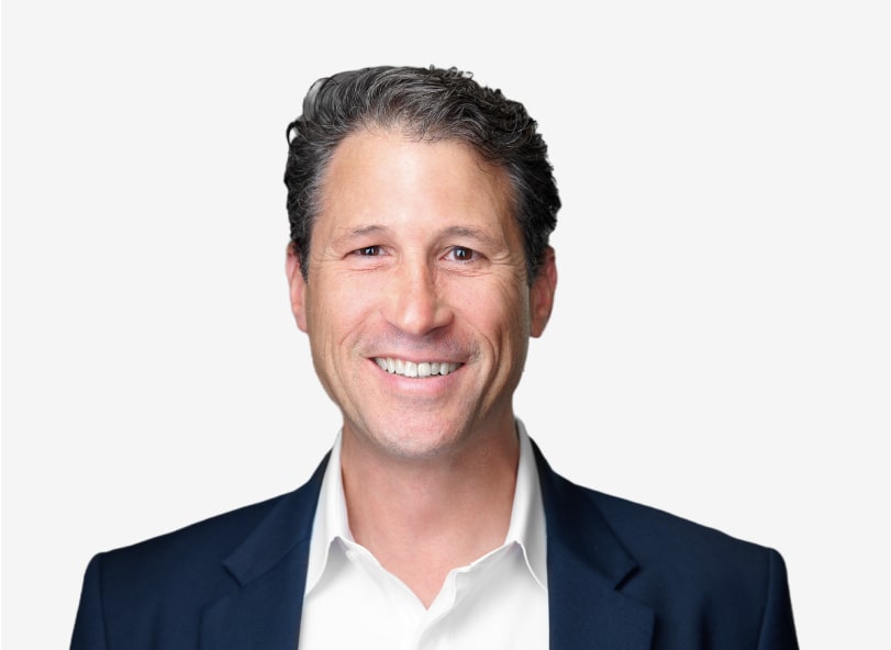 Smiling man with short, wavy dark hair wearing a white dress shirt and dark blazer, posed against a plain white background.