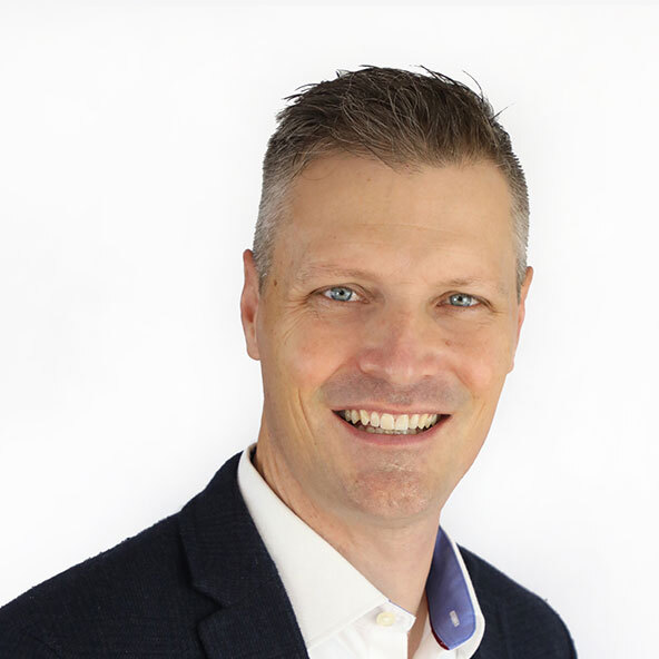 A smiling man with short, light brown hair, wearing a dark blazer over a white collared shirt, poses in front of a plain white background.