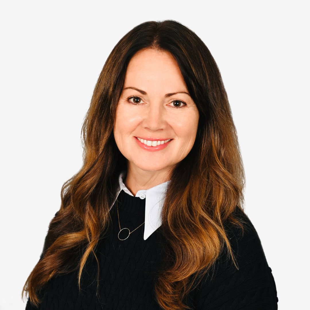 Smiling woman with long brown hair wearing a black sweater over a white collared shirt, posing against a plain white background.