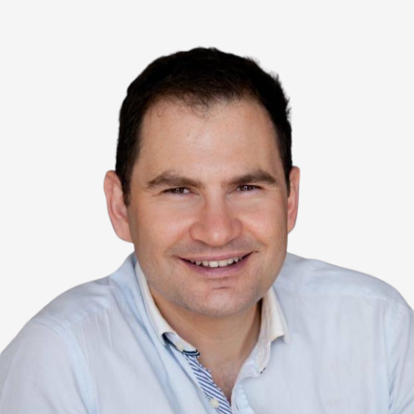 A man with short dark hair, wearing a light blue collared shirt, smiles at the camera against a plain white background.