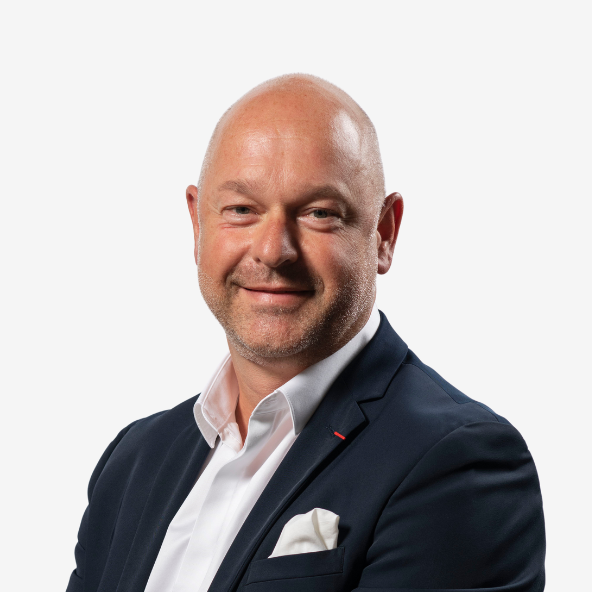 A bald man with a trimmed beard and light complexion, wearing a dark suit jacket, white shirt, and pocket square, smiling confidently against a plain white background.